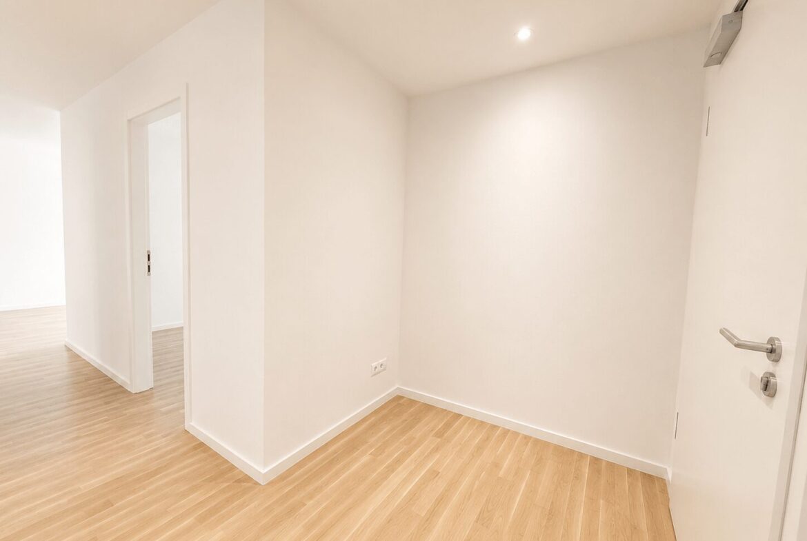Bright, empty hallway with light wood flooring and white walls; a door with a silver handle on the right.