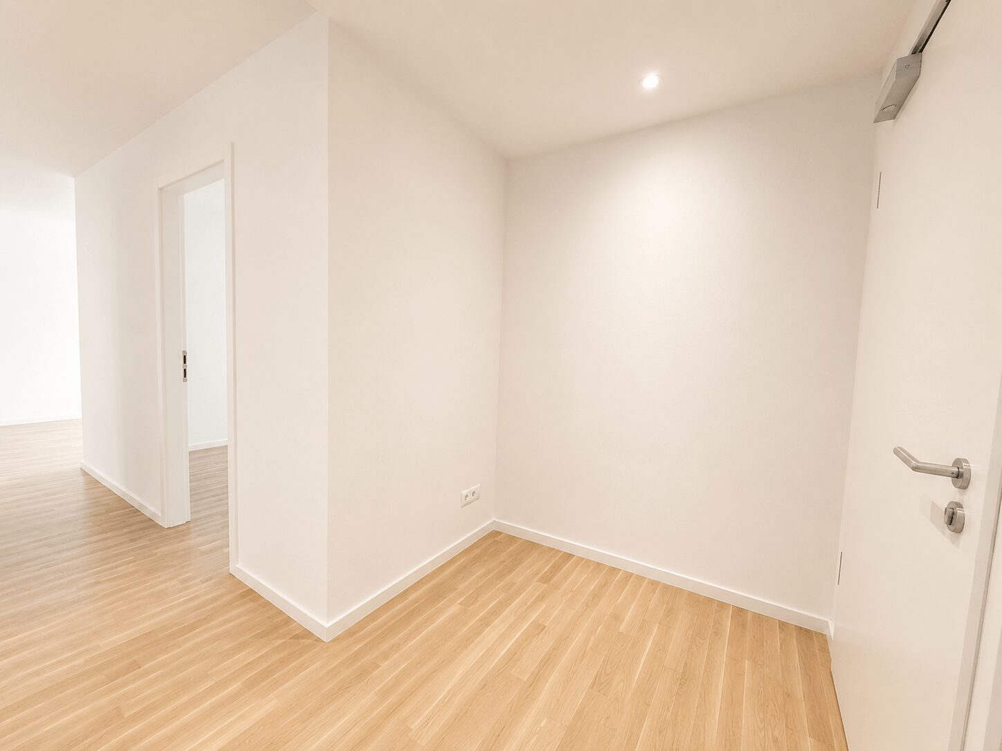 Bright, empty hallway with light wood flooring and white walls; a door with a silver handle on the right.