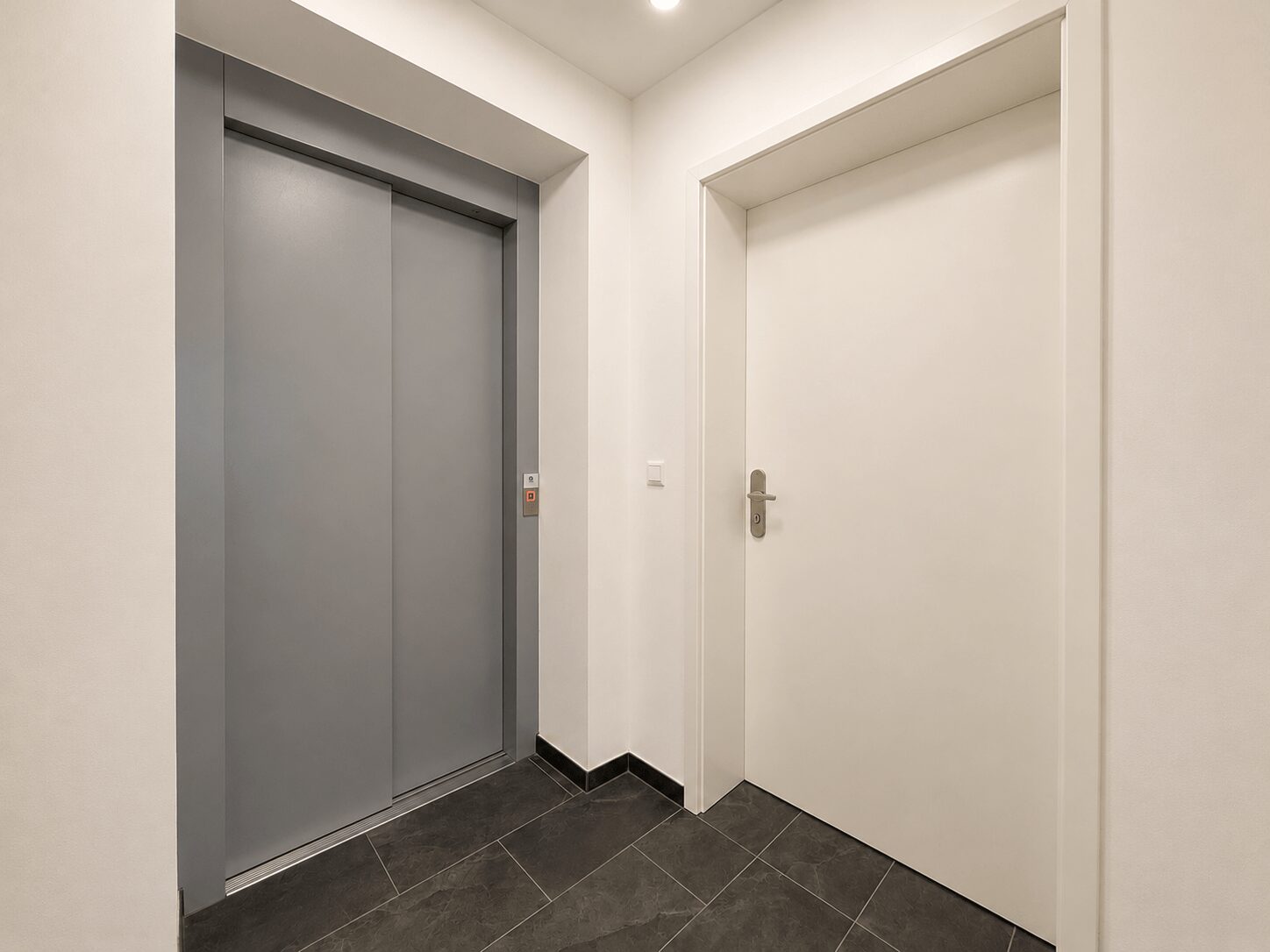 Interior hallway with closed gray elevator doors on the left and a white door on the right, dark tiled floor.