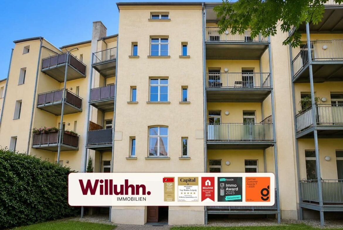 Front view of a beige apartment building with multiple balconies and a large Willuhn Immobilien sign displaying awards in front of the lawn.