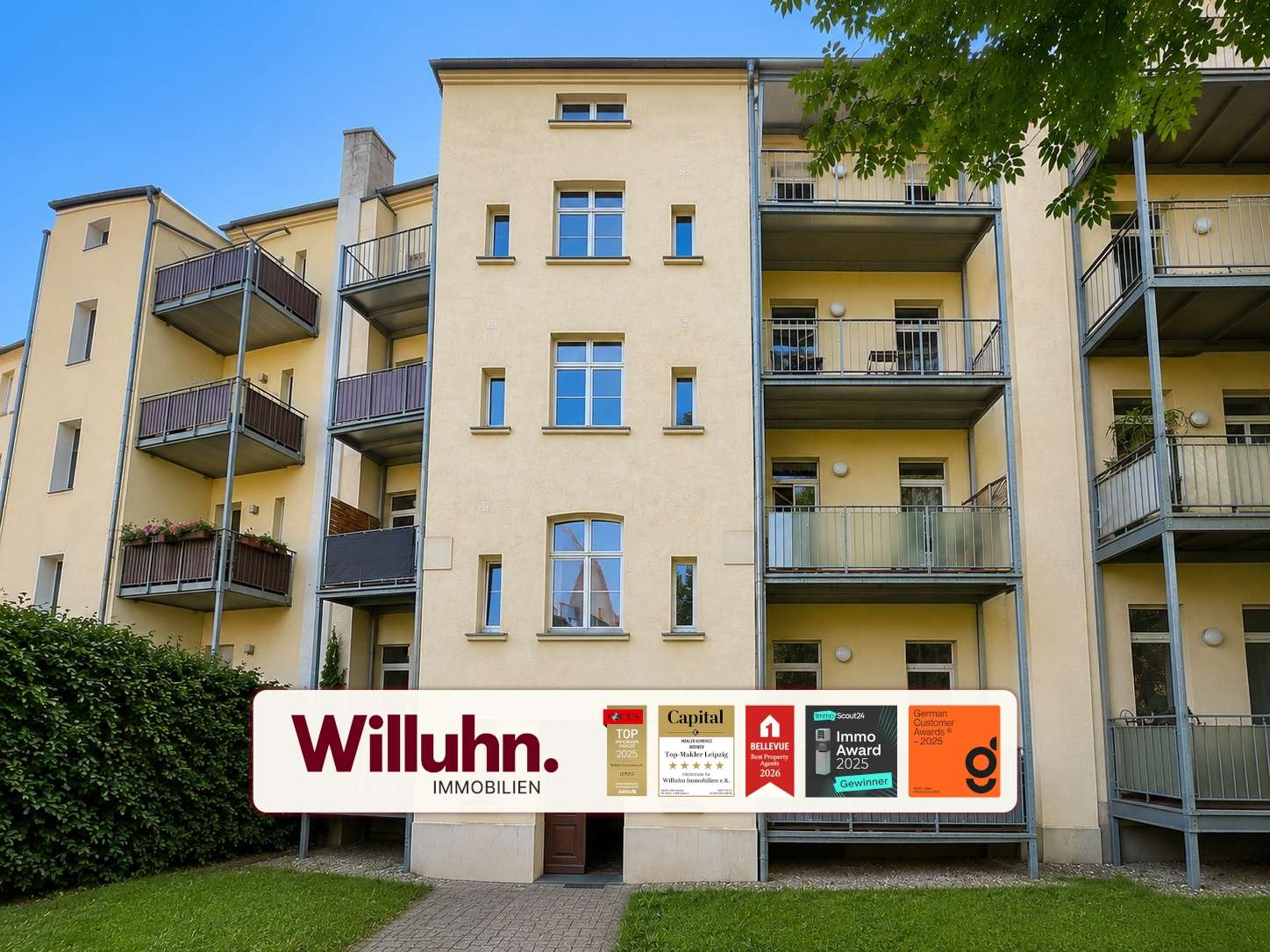 Front view of a beige apartment building with multiple balconies and a large Willuhn Immobilien sign displaying awards in front of the lawn.