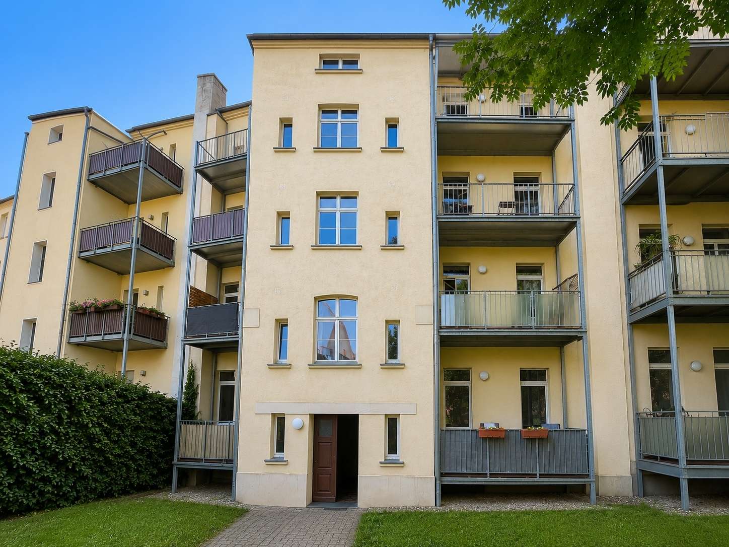 Beige apartment building with multiple balconies and a central entrance under a clear blue sky. A grassy lawn sits in front.