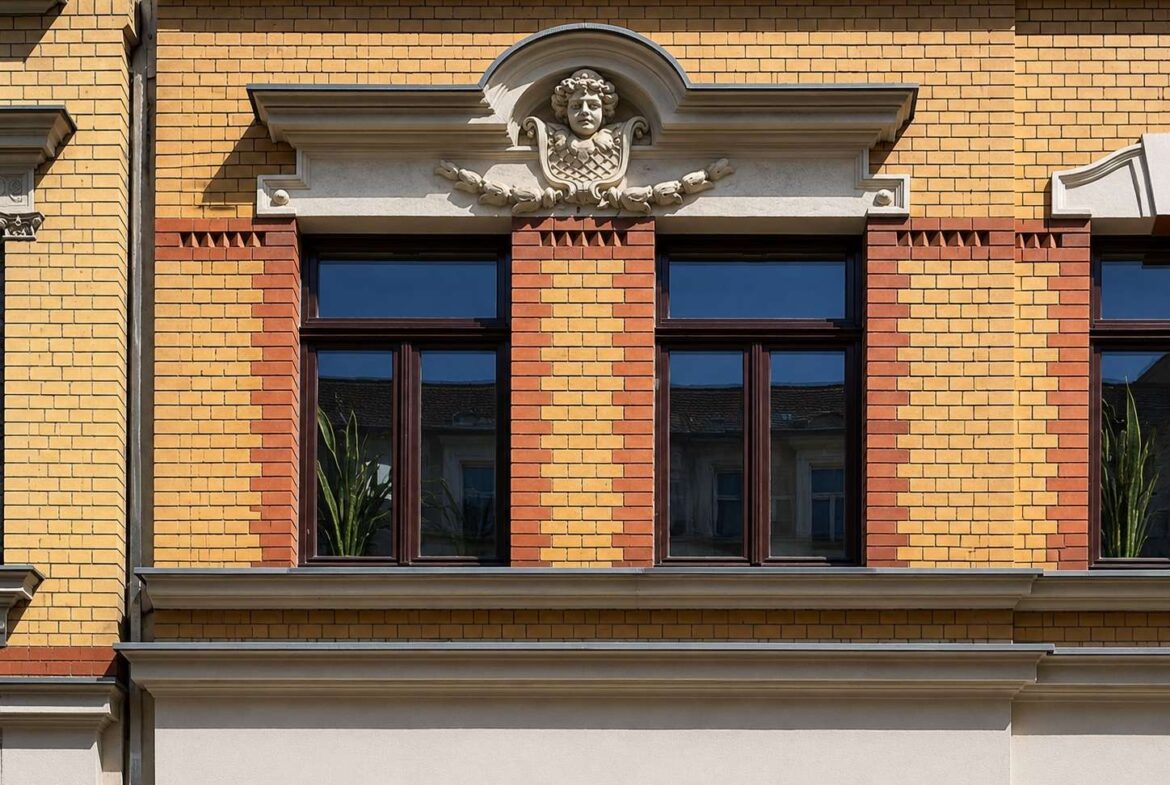 Ornate stone crest with cherub above two tall brick-framed windows on a yellow brick building, with red brick trim surrounding the windows.