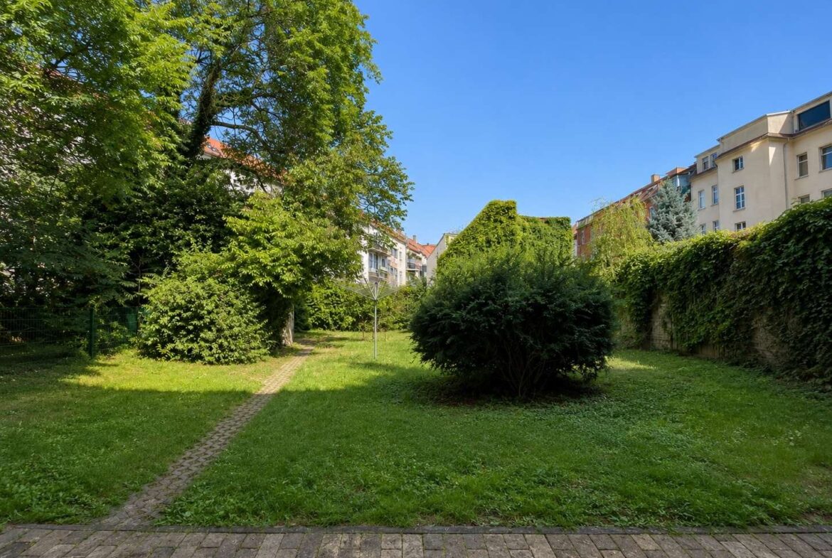 Urban garden courtyard with a paved path, trimmed shrubs, tall trees, and residential buildings in the background.