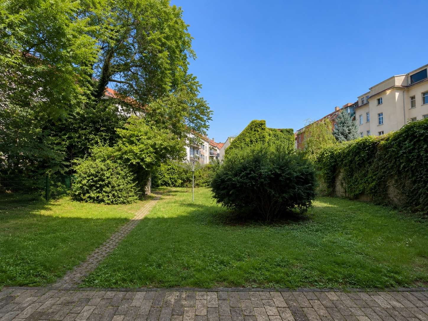 Urban garden courtyard with a paved path, trimmed shrubs, tall trees, and residential buildings in the background.