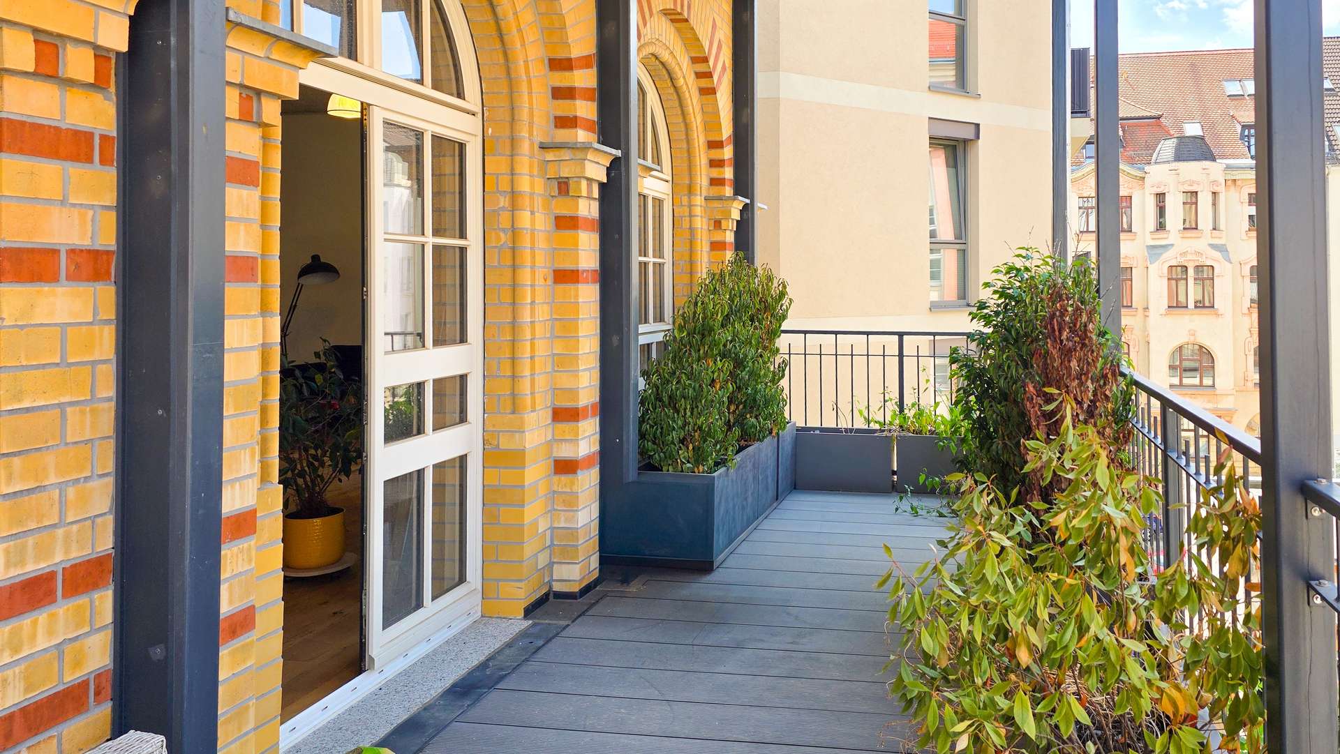 Balcony with potted shrubs along a blue railing outside a yellow brick building; open glass French doors.