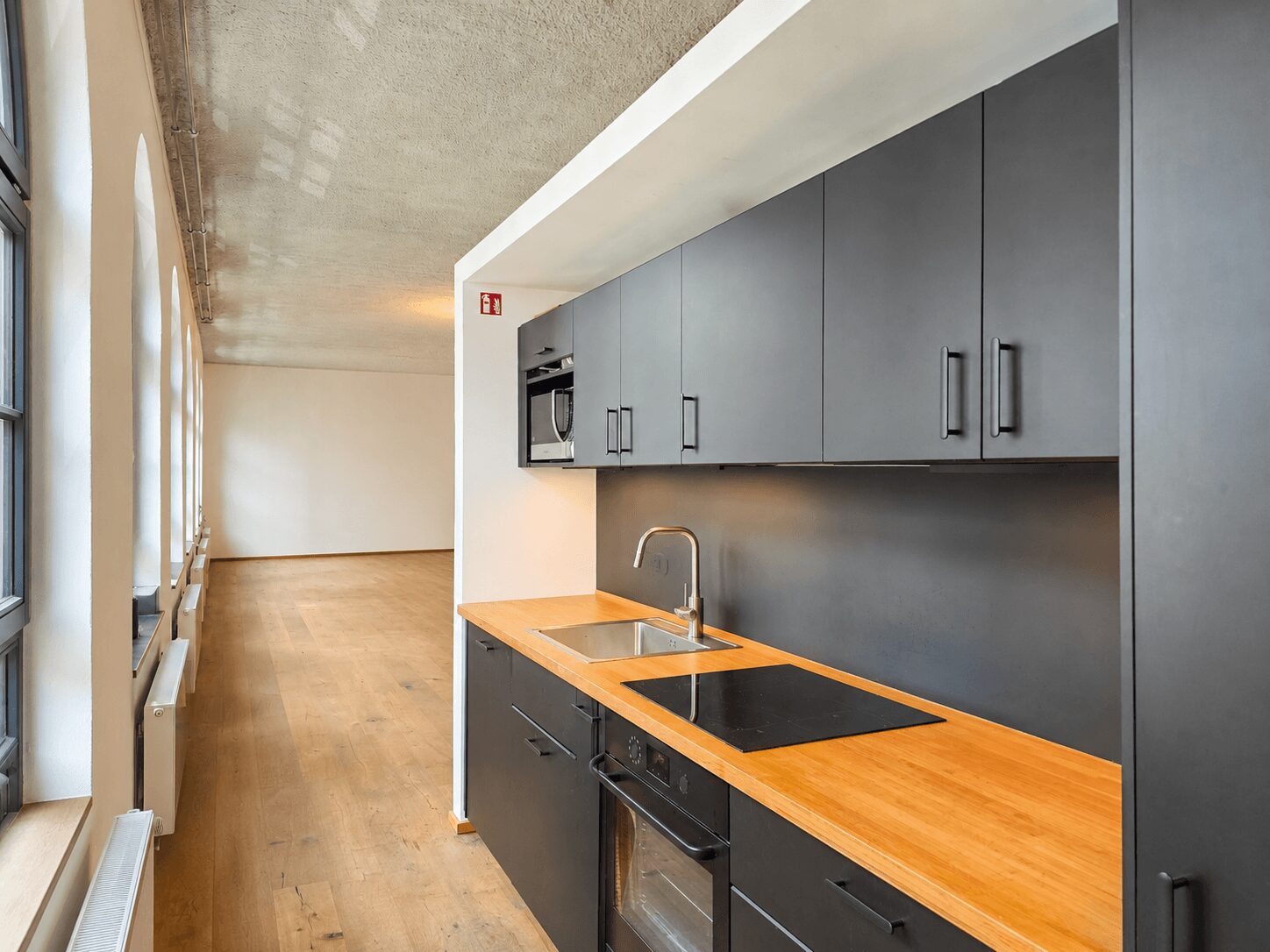 Narrow modern kitchen with matte black cabinets, wood countertop, sink, and induction hob; sunny windows along the left wall.