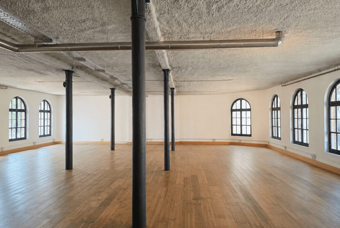 Empty airy studio with arched windows, light wood floor, and black metal columns.