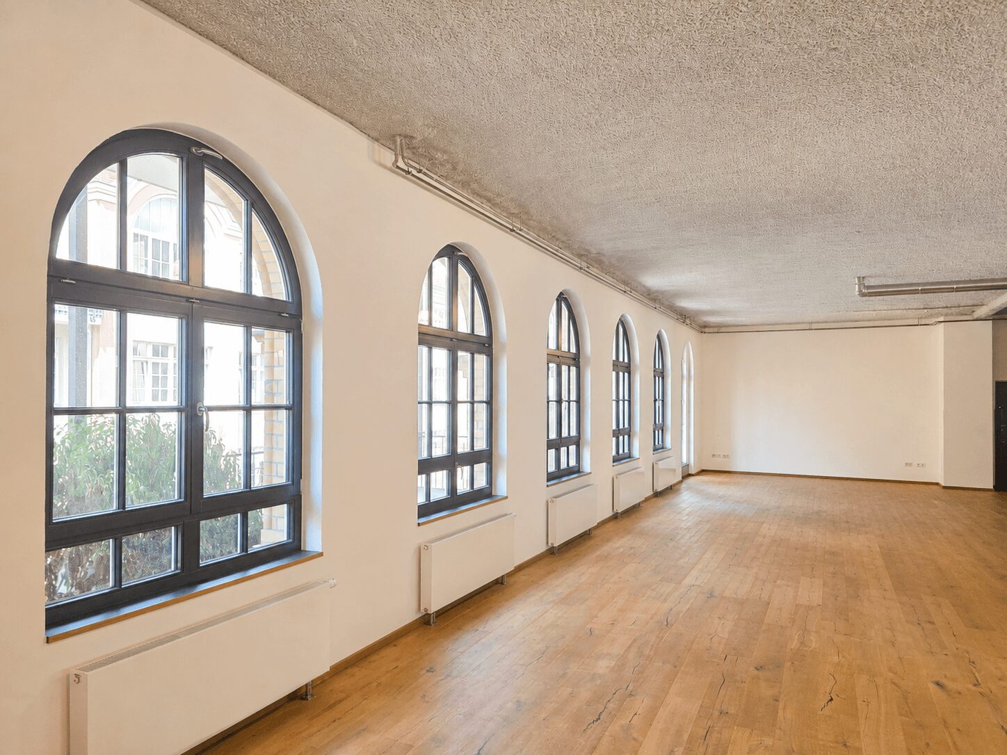 Bright empty room with a row of arched windows along one wall, radiators under each window, and a light wooden floor.