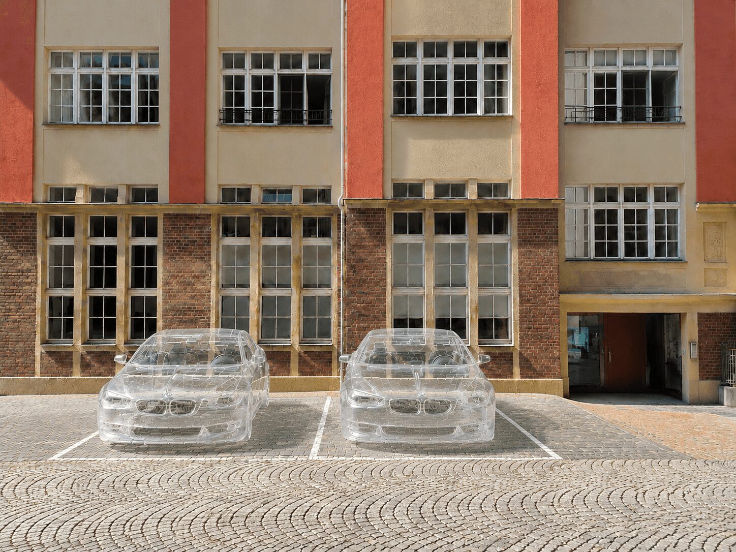 Transparent car sculptures parked in front of a brick-and-beige building in a cobblestone lot