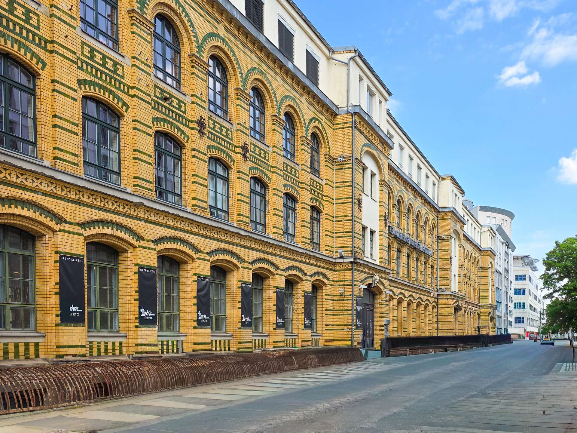 Yellow brick building with green decorative arches and large arched windows along a quiet street. (Informative)