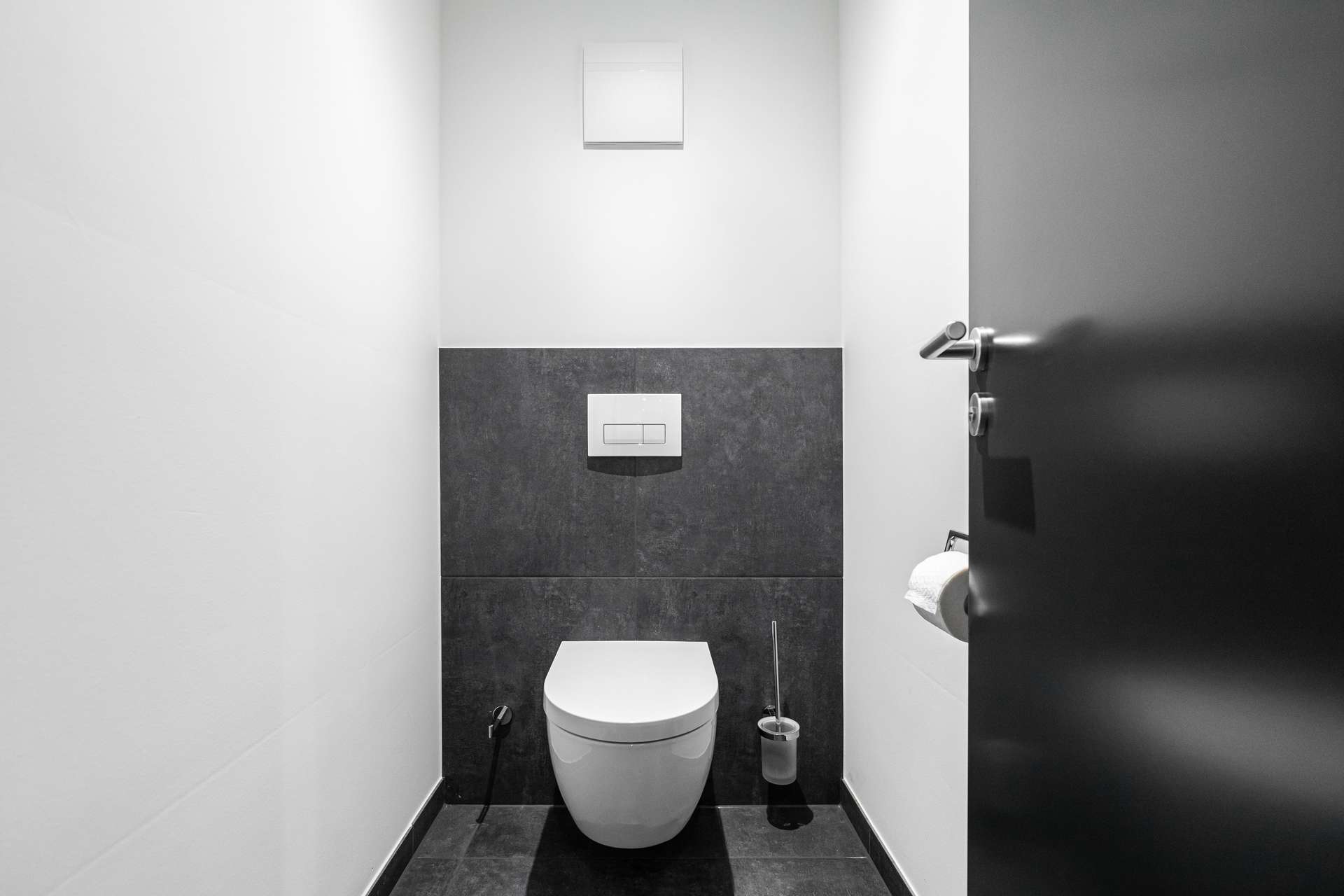 Compact modern bathroom with a wall-hung toilet and dual-flush plate against a dark tiled wall; door is partly open to the right.