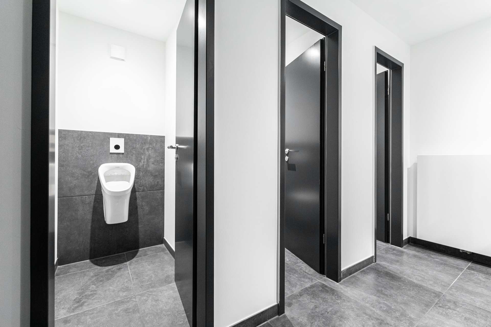 Public restroom hallway with three stalls and a wall-mounted urinal visible in the left stall, white walls and dark doorway frames.