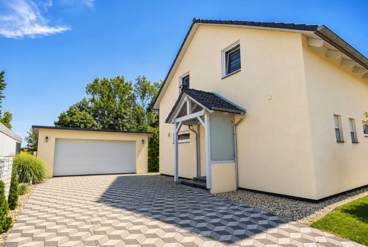 Beige house with a two-car garage, patterned geometric driveway, and small front porch under a blue sky.