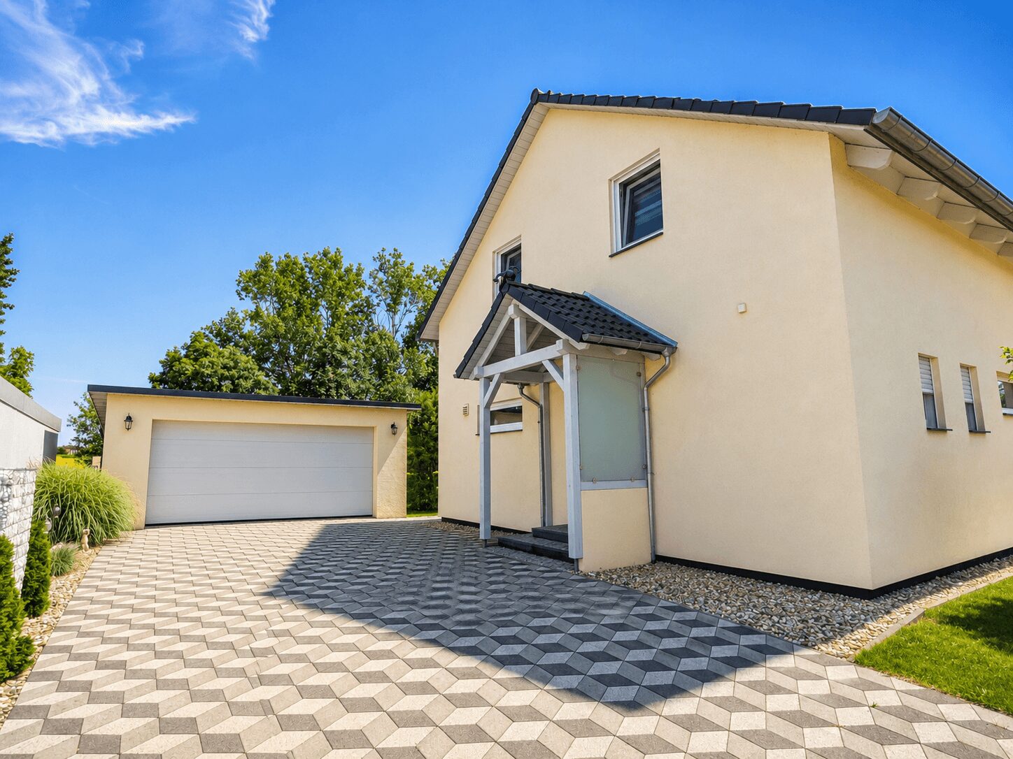 Beige house with a two-car garage, patterned geometric driveway, and small front porch under a blue sky.