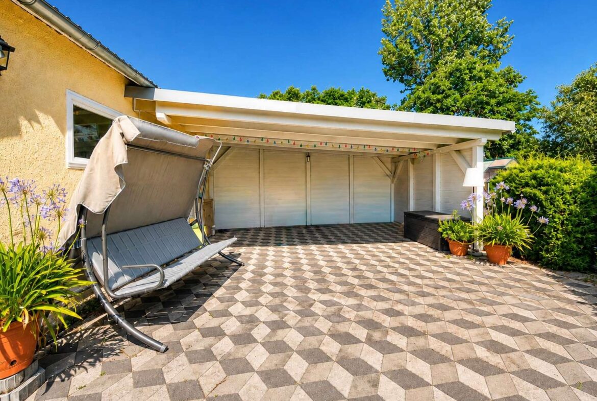 Sunny backyard patio with geometric tile floor, a gray outdoor swing, potted plants, and a white covered structure against a yellow house wall.