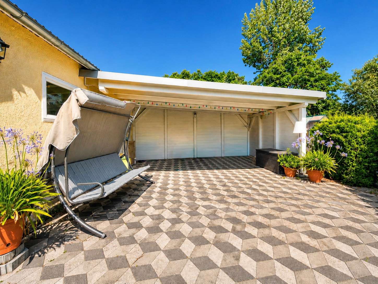 Sunny backyard patio with geometric tile floor, a gray outdoor swing, potted plants, and a white covered structure against a yellow house wall.