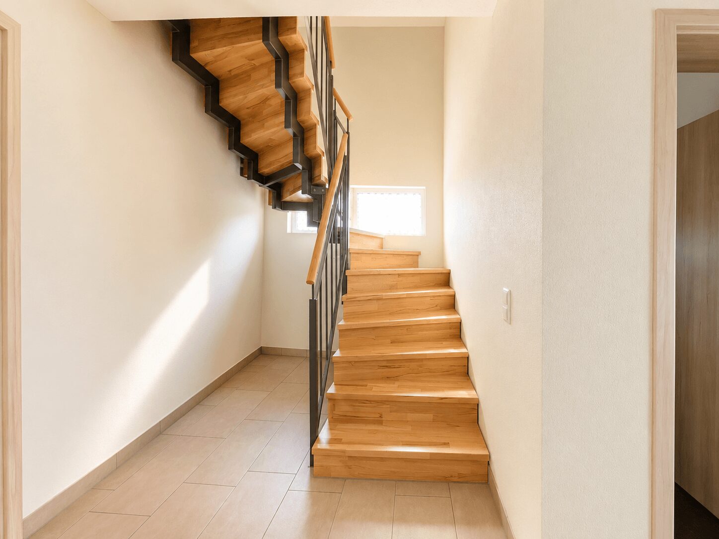 Wooden stairs with a metal railing leading to an upper floor in a bright beige hallway, illuminated by a window on the landing.