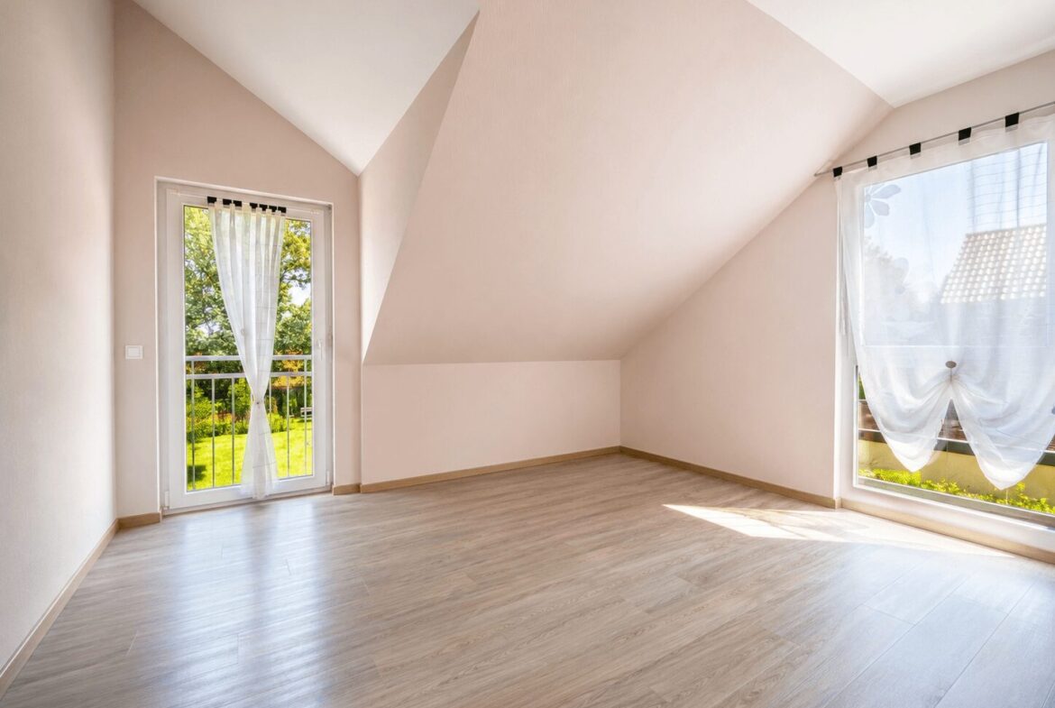 Bright empty attic room with sloped walls, light wood flooring, and a glass door with sheer curtain leading to a balcony.
