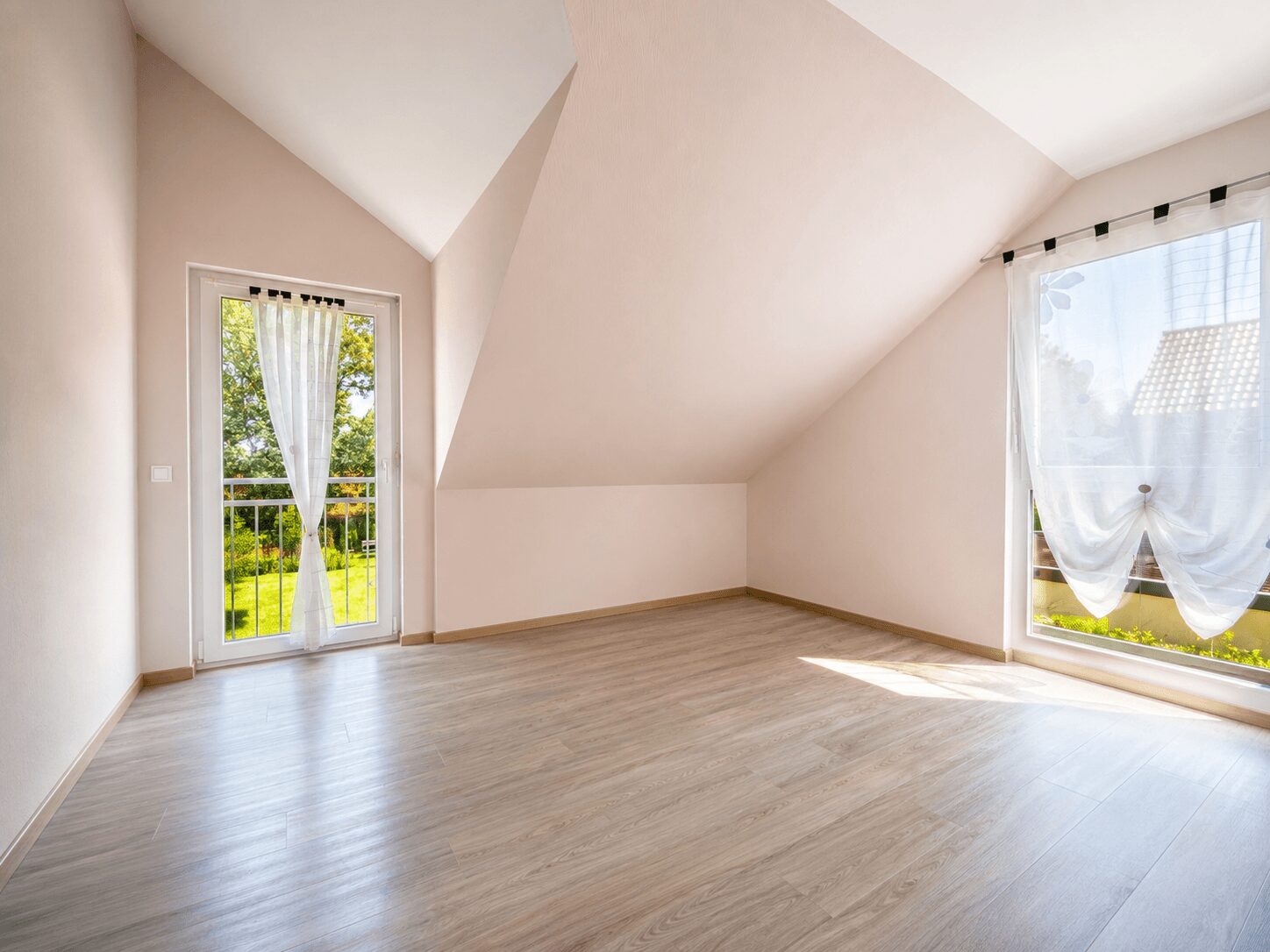 Bright empty attic room with sloped walls, light wood flooring, and a glass door with sheer curtain leading to a balcony.