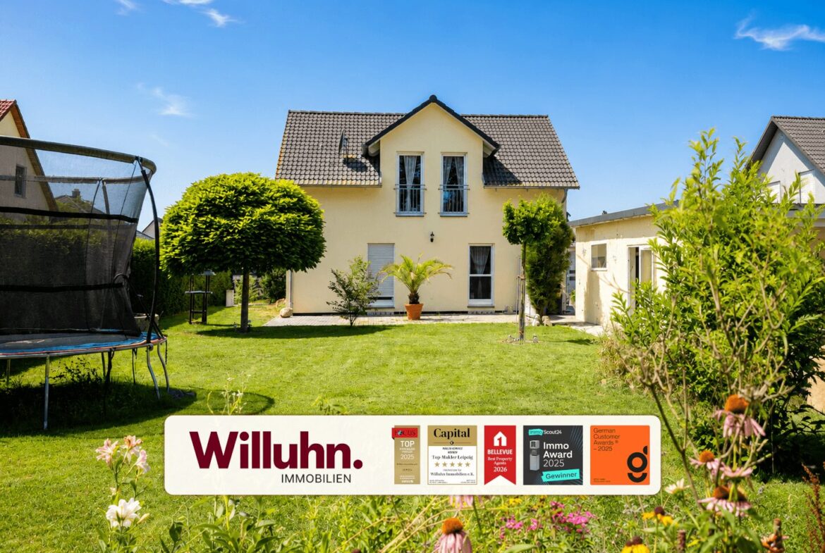 Two-story yellow house with a well-kept lawn and a trampoline on the left, under a clear blue sky; real estate sign in the foreground reads Willuhn Immobilien with awards icons nearby.
