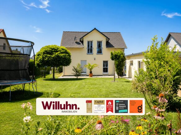Two-story yellow house with a well-kept lawn and a trampoline on the left, under a clear blue sky; real estate sign in the foreground reads Willuhn Immobilien with awards icons nearby.