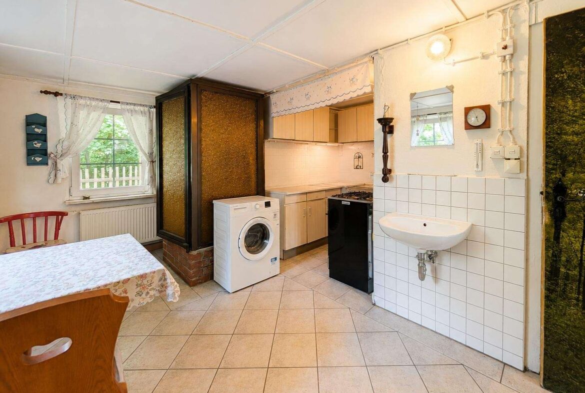 Cozy kitchen with a washing machine, gas stove, and sink along a tiled wall, and a small dining table by a window with lace curtains.