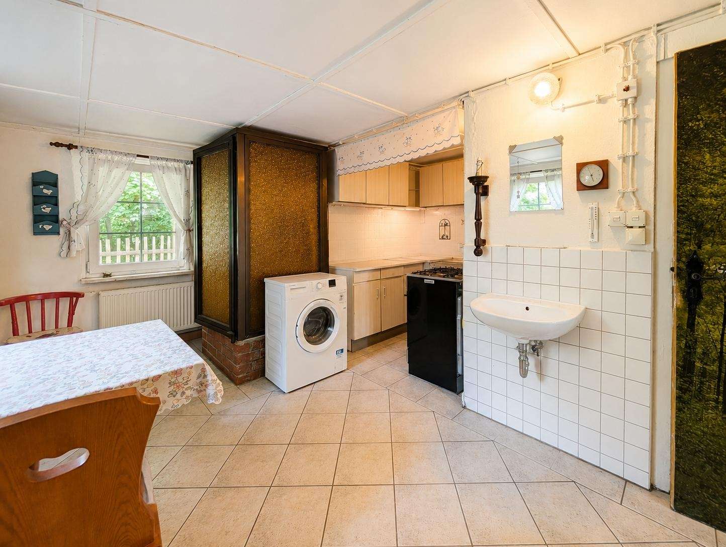Cozy kitchen with a washing machine, gas stove, and sink along a tiled wall, and a small dining table by a window with lace curtains.