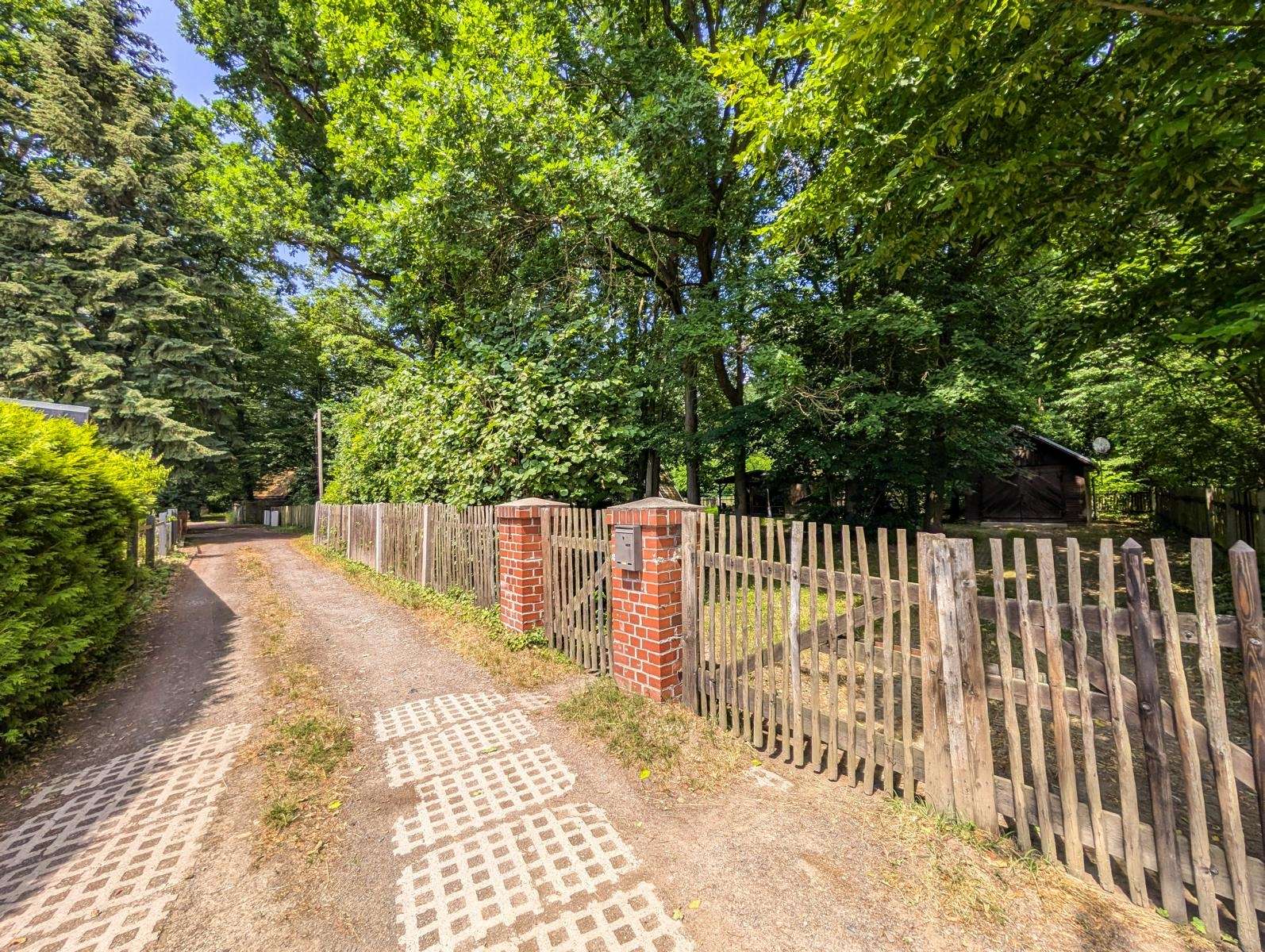 Dirt path lined with wooden fence and brick gate pillars, surrounded by dense green trees and a small shed in the background.