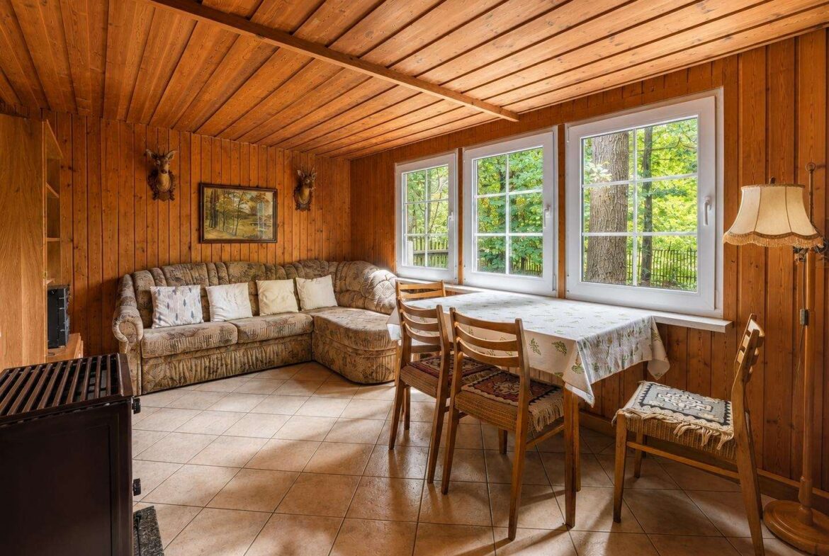 Rustic living-dining area with wood paneled walls and ceiling, an L-shaped sofa, and a table by three windows showing greenery outside.