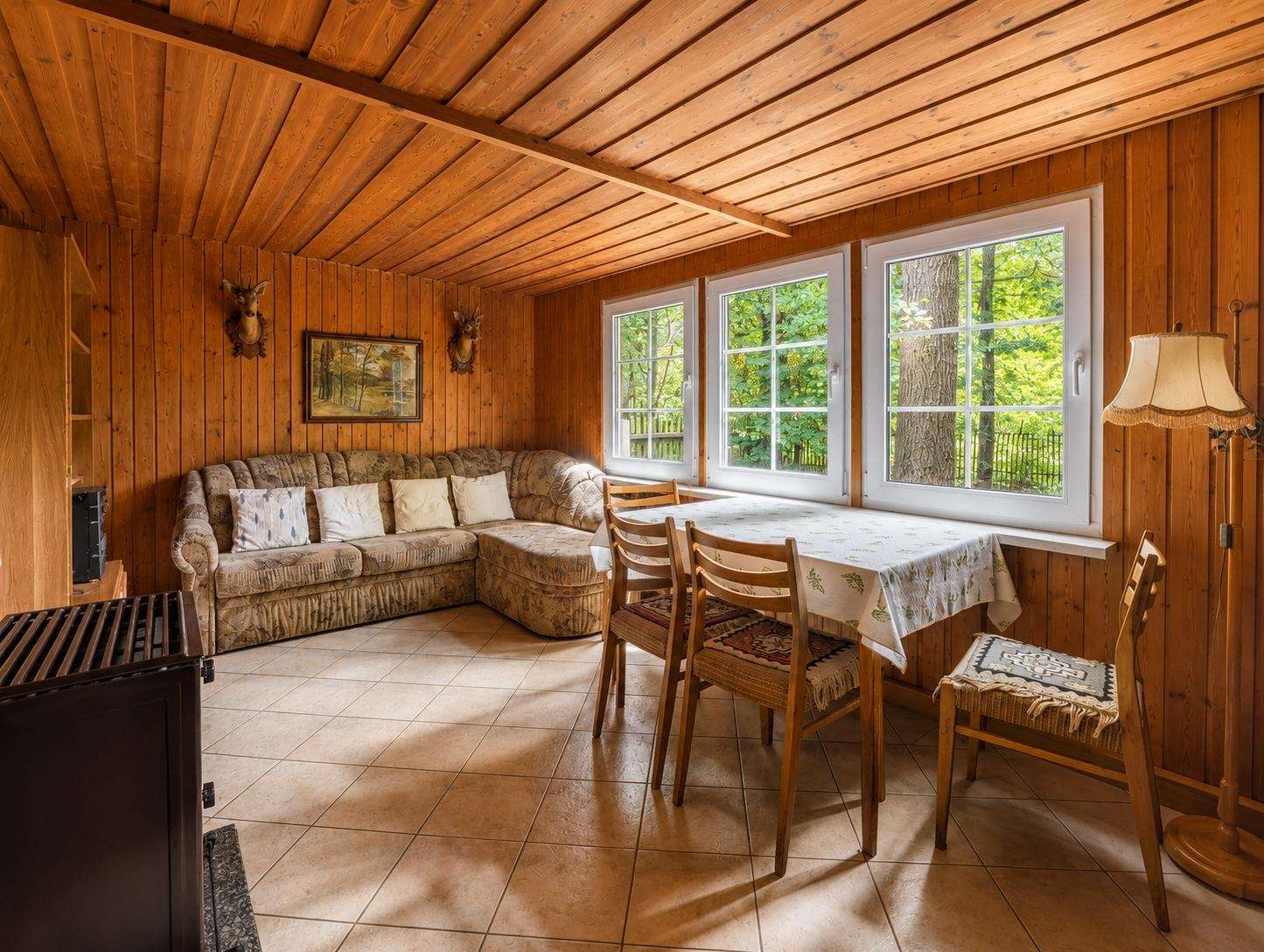 Rustic living-dining area with wood paneled walls and ceiling, an L-shaped sofa, and a table by three windows showing greenery outside.