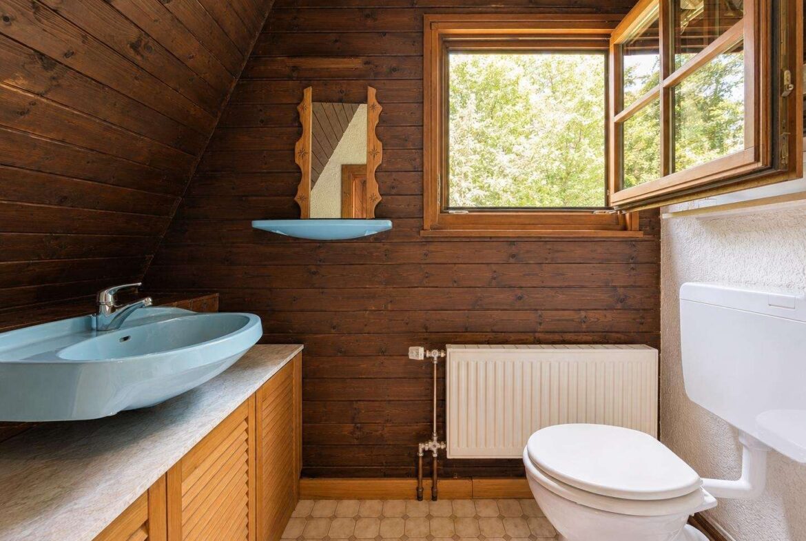 Rustic bathroom with dark wood paneling, a blue oval sink on a Marble-like countertop, and a window overlooking greenery outside.
