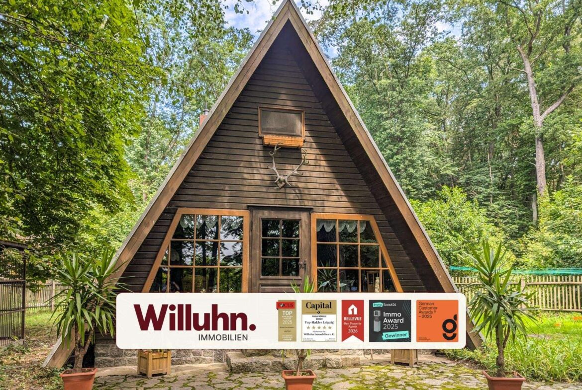 A dark brown A-frame wooden cabin in a forest, with a large real estate sign (Willuhn Immobilien) in front.