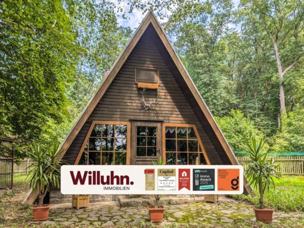 A dark brown A-frame wooden cabin in a forest, with a large real estate sign (Willuhn Immobilien) in front.