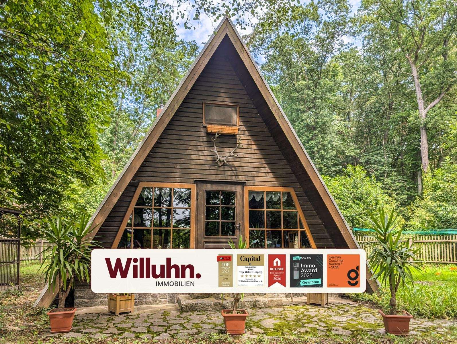 A dark brown A-frame wooden cabin in a forest, with a large real estate sign (Willuhn Immobilien) in front.