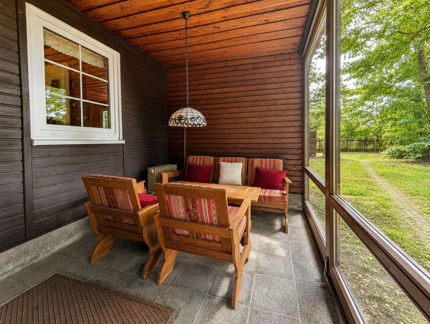 Wooden-paneled sunroom with a striped-cushion seating set around a wooden coffee table, large windows to a green yard, and a hanging Tiffany-style lamp above.