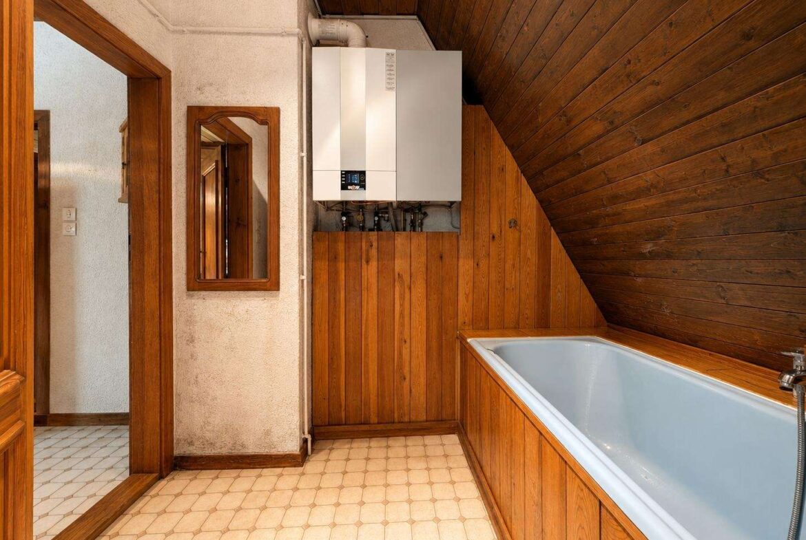 A bathroom with a white boiler mounted on a wooden paneled wall, next to a rectangular mirror and wooden doorframe; tiled floor and a corner bathtub with wooden paneling along the walls under a sloped wooden ceiling.