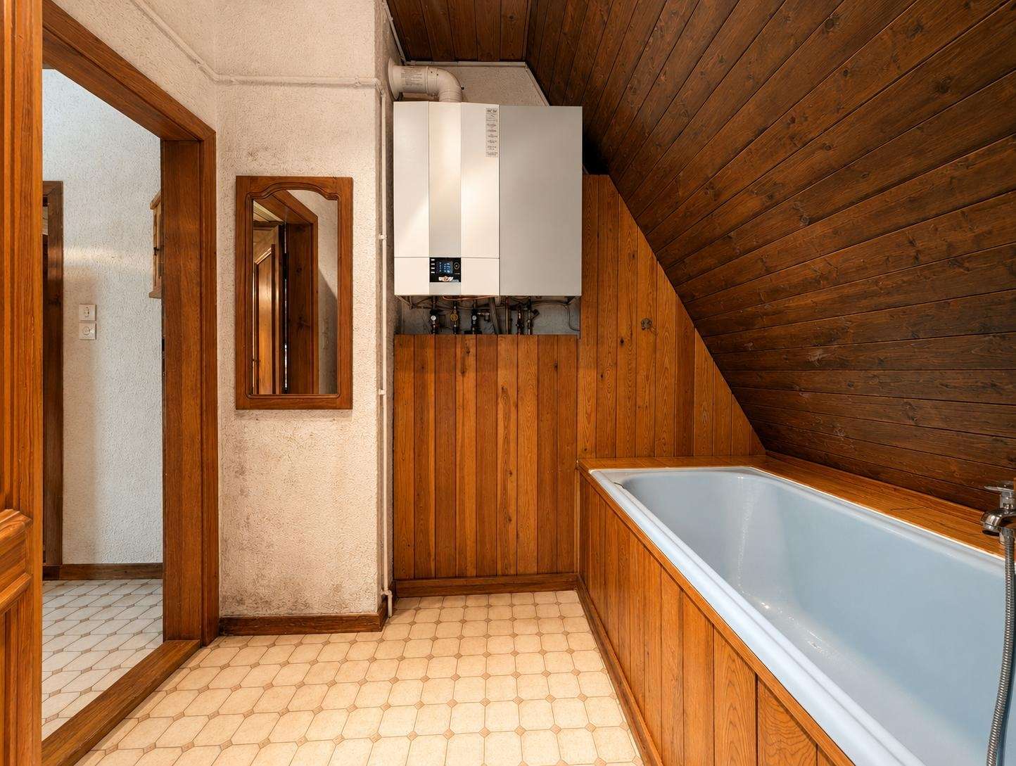 A bathroom with a white boiler mounted on a wooden paneled wall, next to a rectangular mirror and wooden doorframe; tiled floor and a corner bathtub with wooden paneling along the walls under a sloped wooden ceiling.