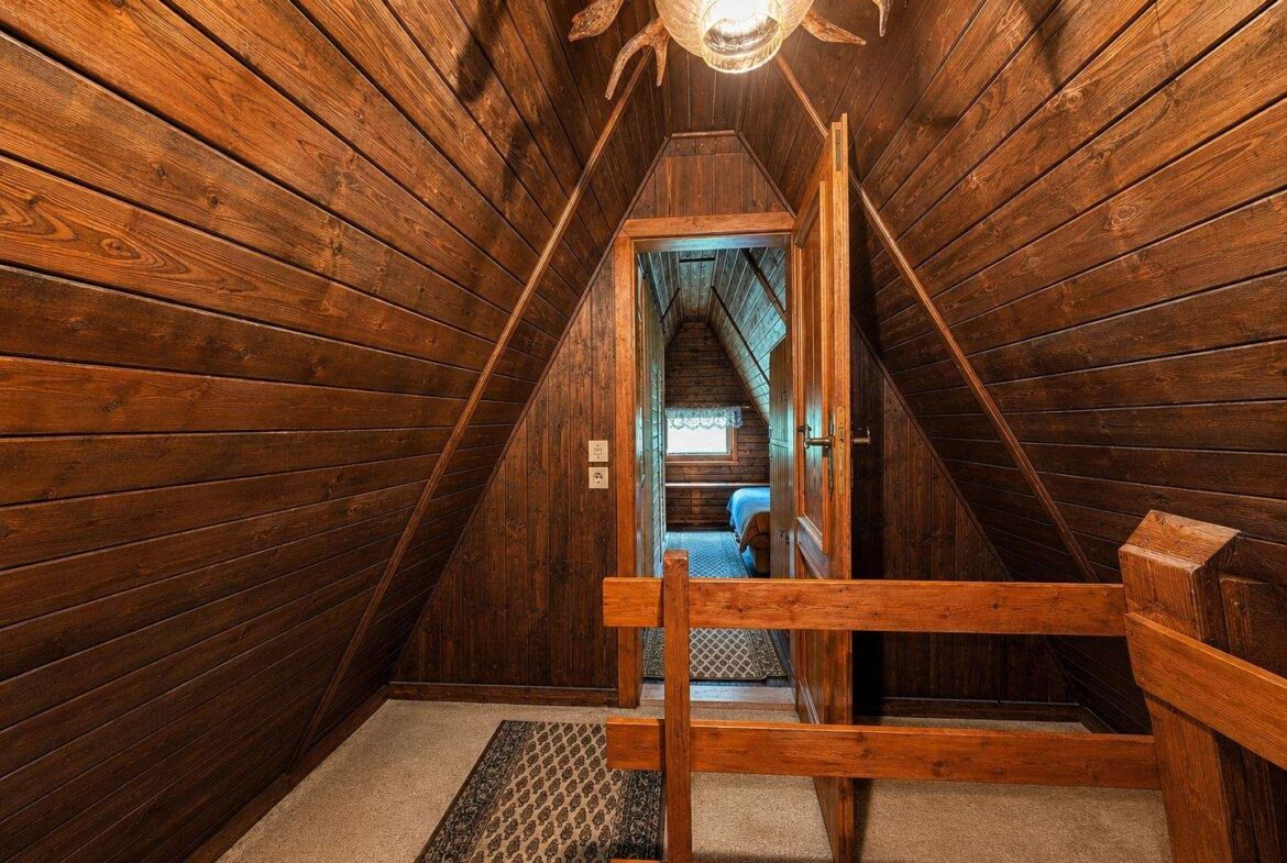 Wood-paneled attic hallway with sloped walls, a wooden railing, and a door opening to a small bedroom at the end; warm light from a ceiling fixture overhead.
