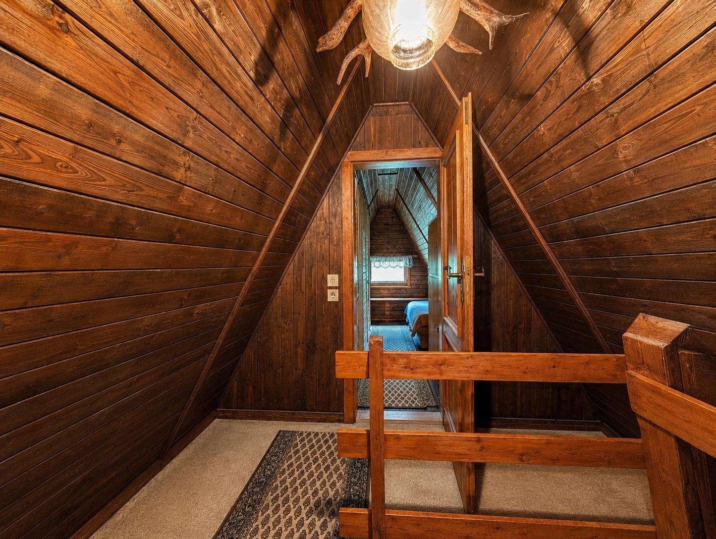 Wood-paneled attic hallway with sloped walls, a wooden railing, and a door opening to a small bedroom at the end; warm light from a ceiling fixture overhead.