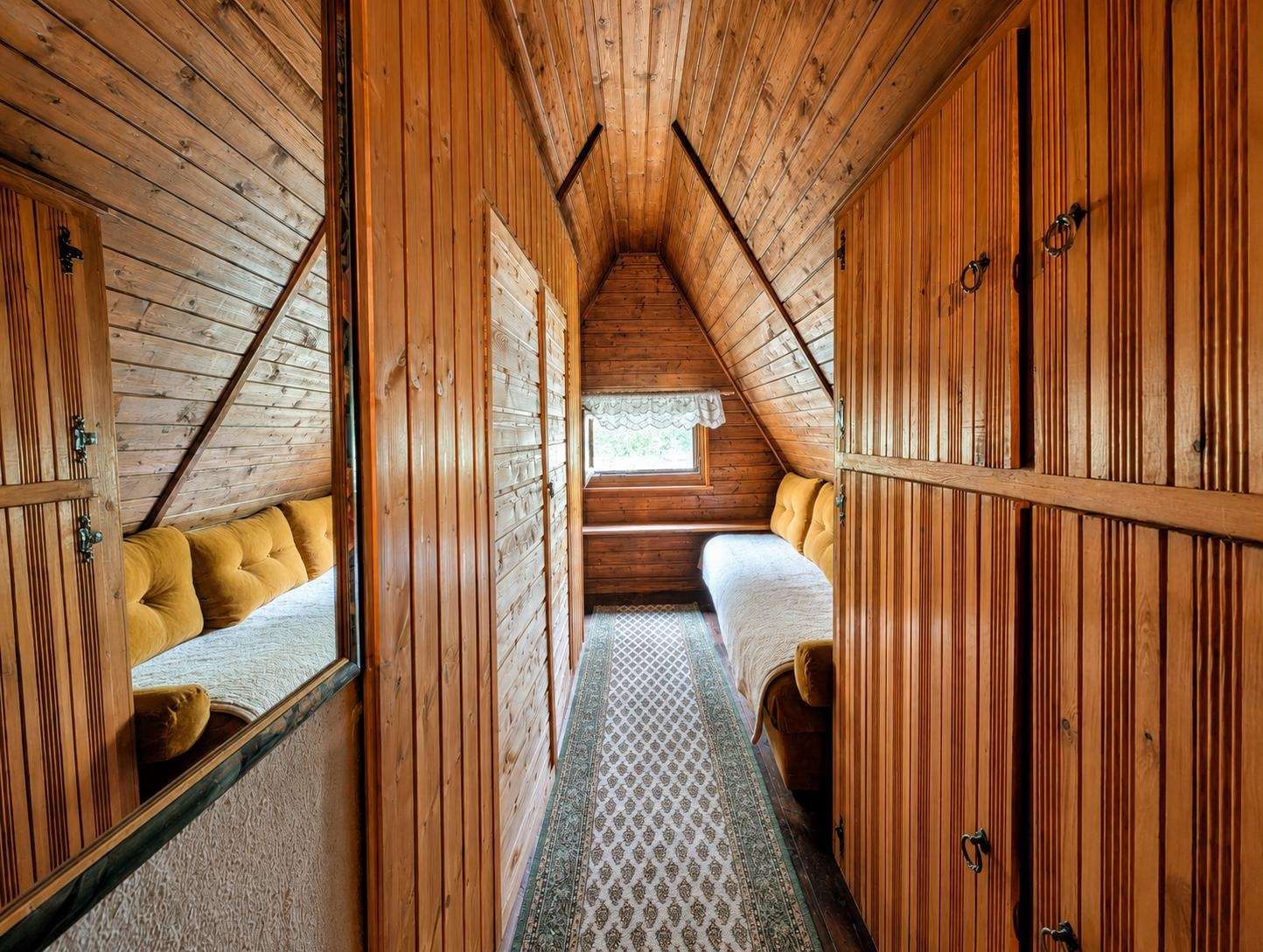 Rustic wooden attic bedroom with two single beds along opposite walls, mustard cushions, and a window at the far end.