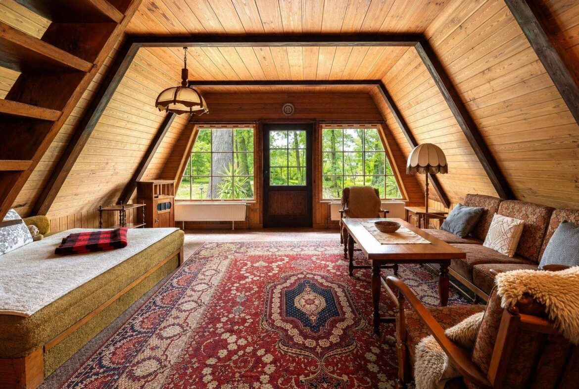 Cozy attic living room with wood paneling, large windows, and a red patterned rug; sofa, chairs, and a wooden coffee table.