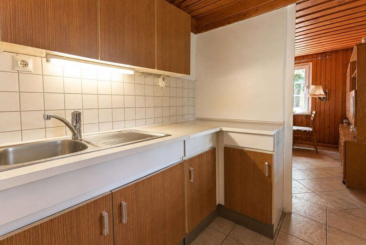 Kitchen with brown wooden cabinets, white countertops, and tiled backsplash; doorway opens to a wood-paneled living area.