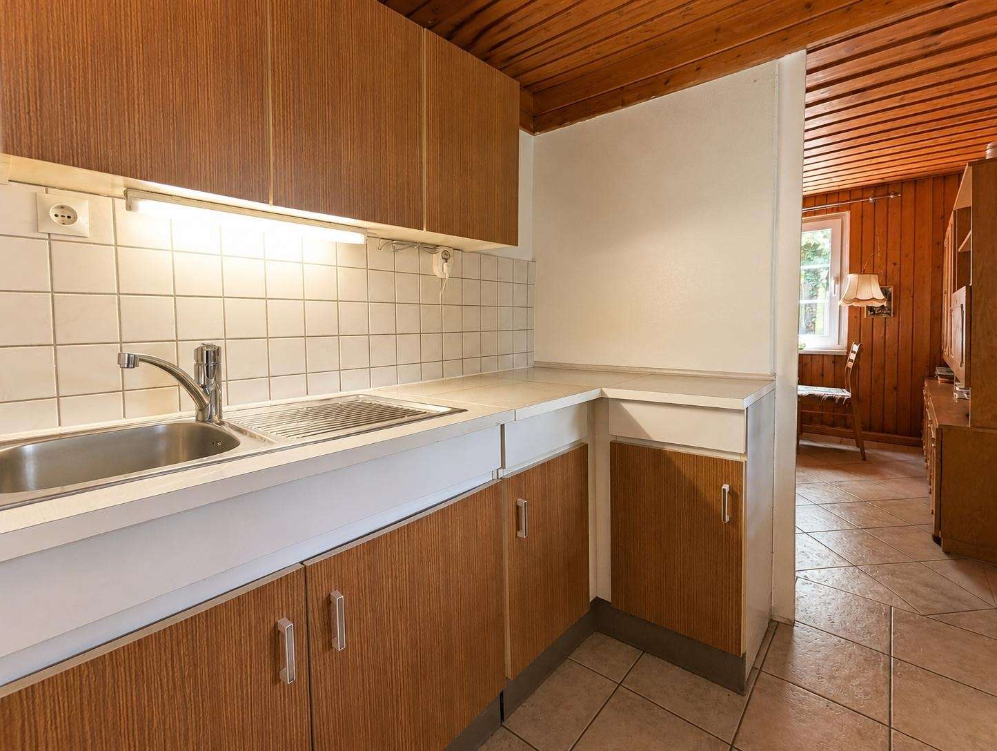 Kitchen with brown wooden cabinets, white countertops, and tiled backsplash; doorway opens to a wood-paneled living area.