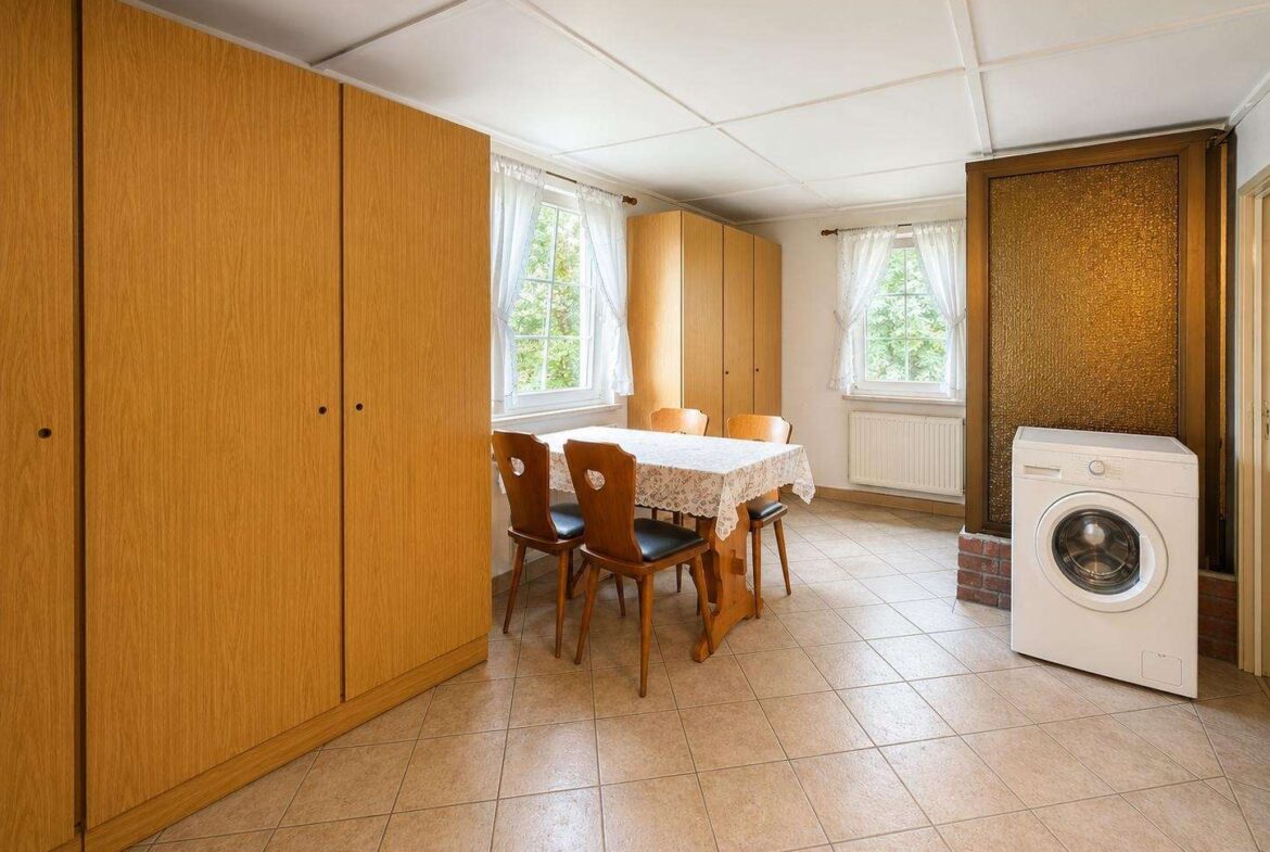 A bright dining area with a wooden table and four chairs, lace tablecloth, and tall wooden cabinets along the wall next to two windows with sheer curtains.