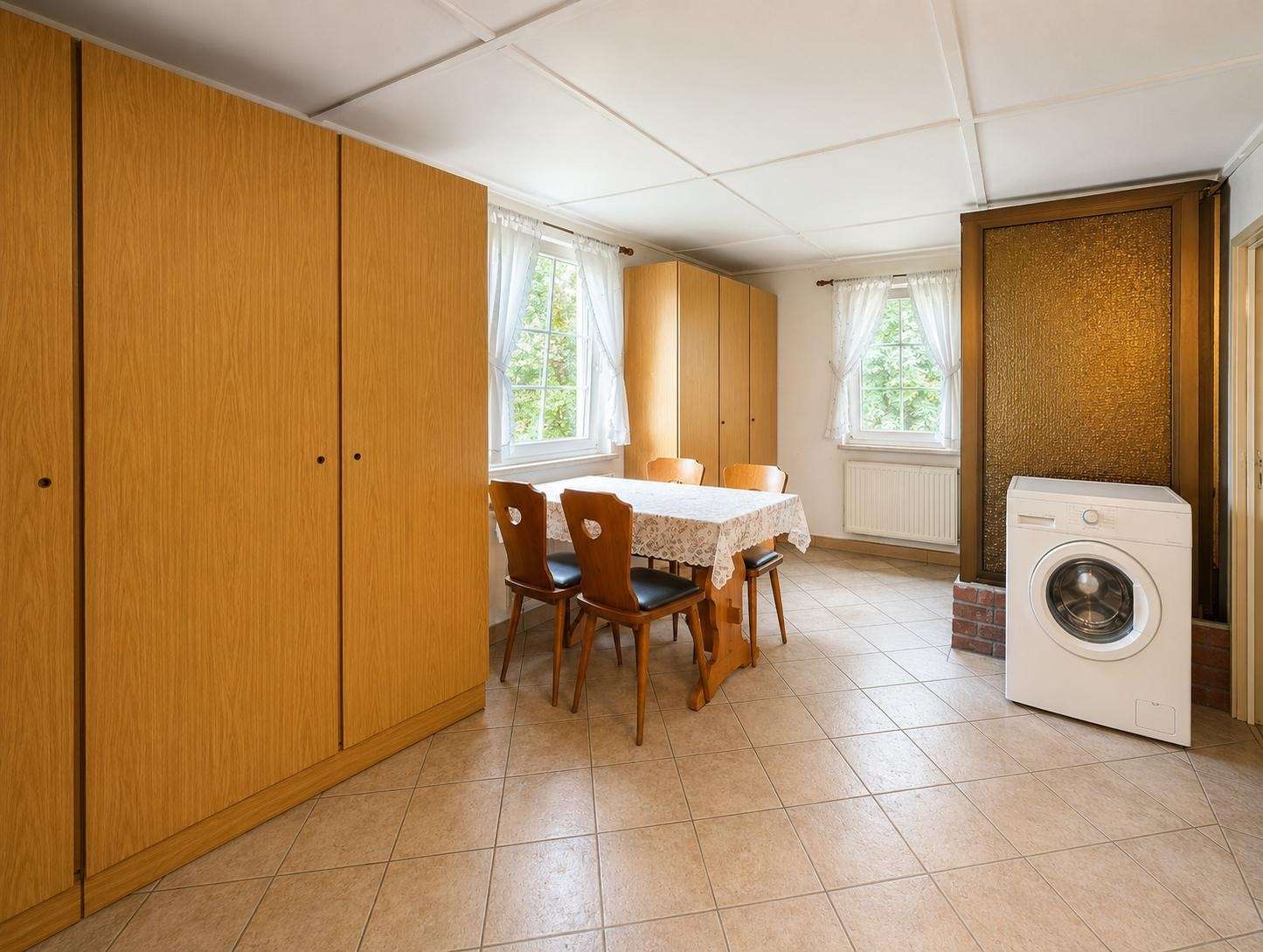 A bright dining area with a wooden table and four chairs, lace tablecloth, and tall wooden cabinets along the wall next to two windows with sheer curtains.
