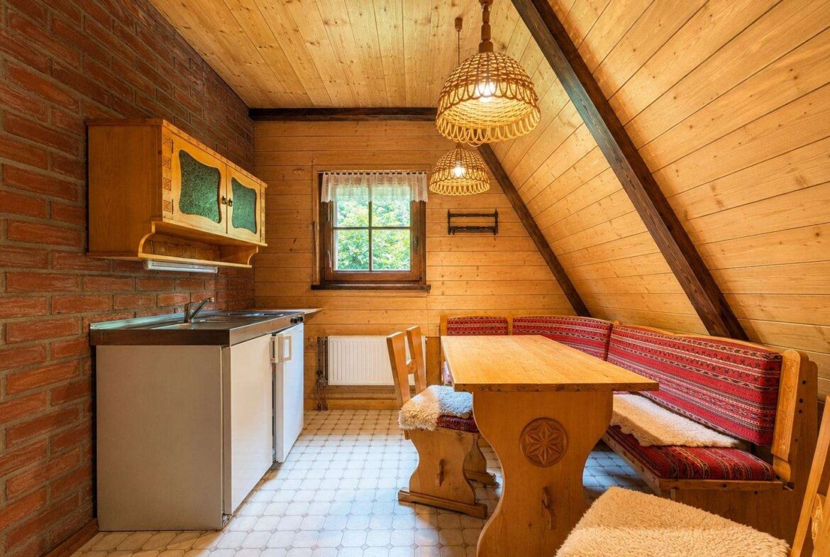 Rustic attic kitchenette with brick wall, wooden cabinets, sink, mini fridge, and a dining nook with red patterned benches.