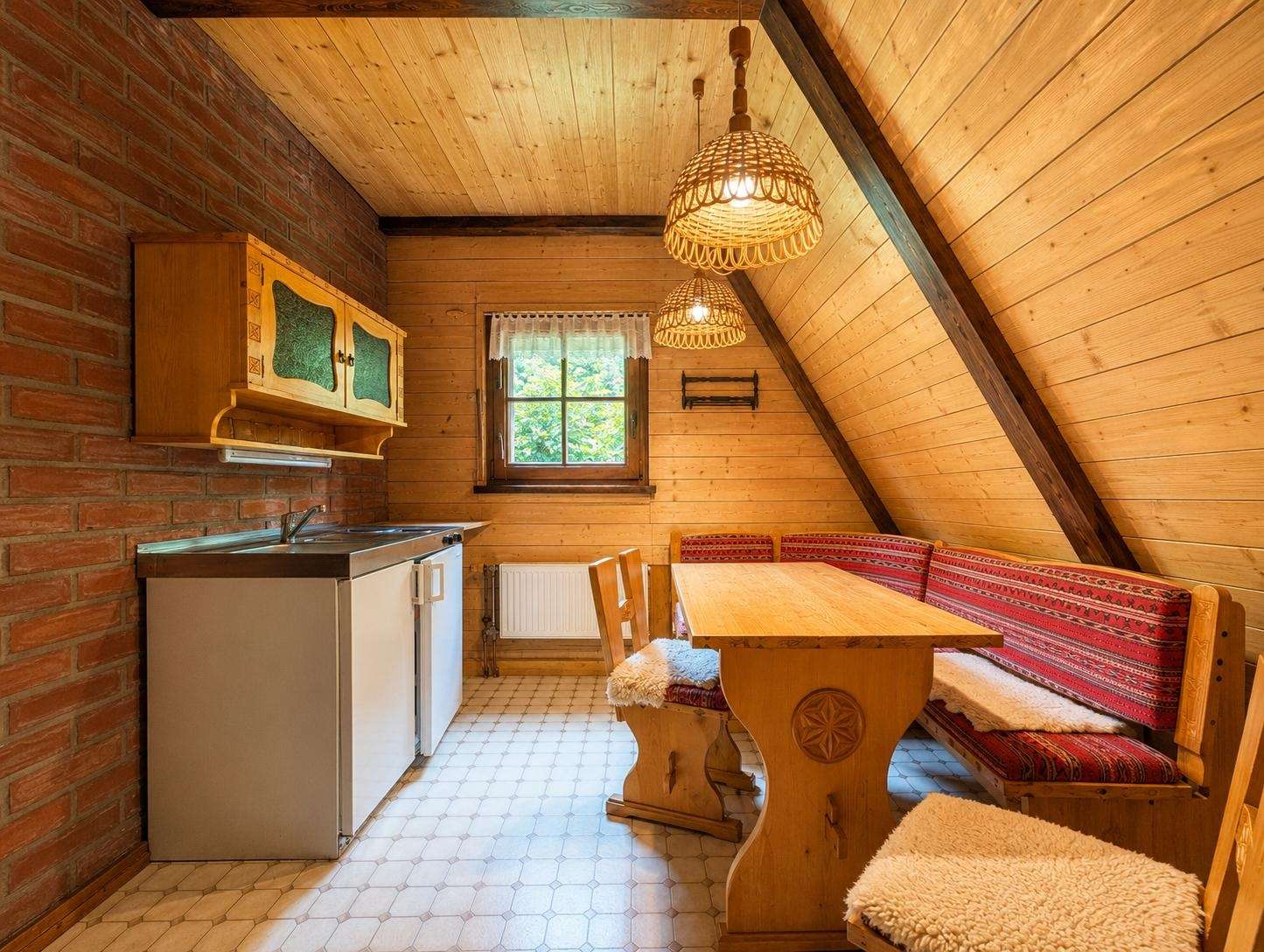 Rustic attic kitchenette with brick wall, wooden cabinets, sink, mini fridge, and a dining nook with red patterned benches.