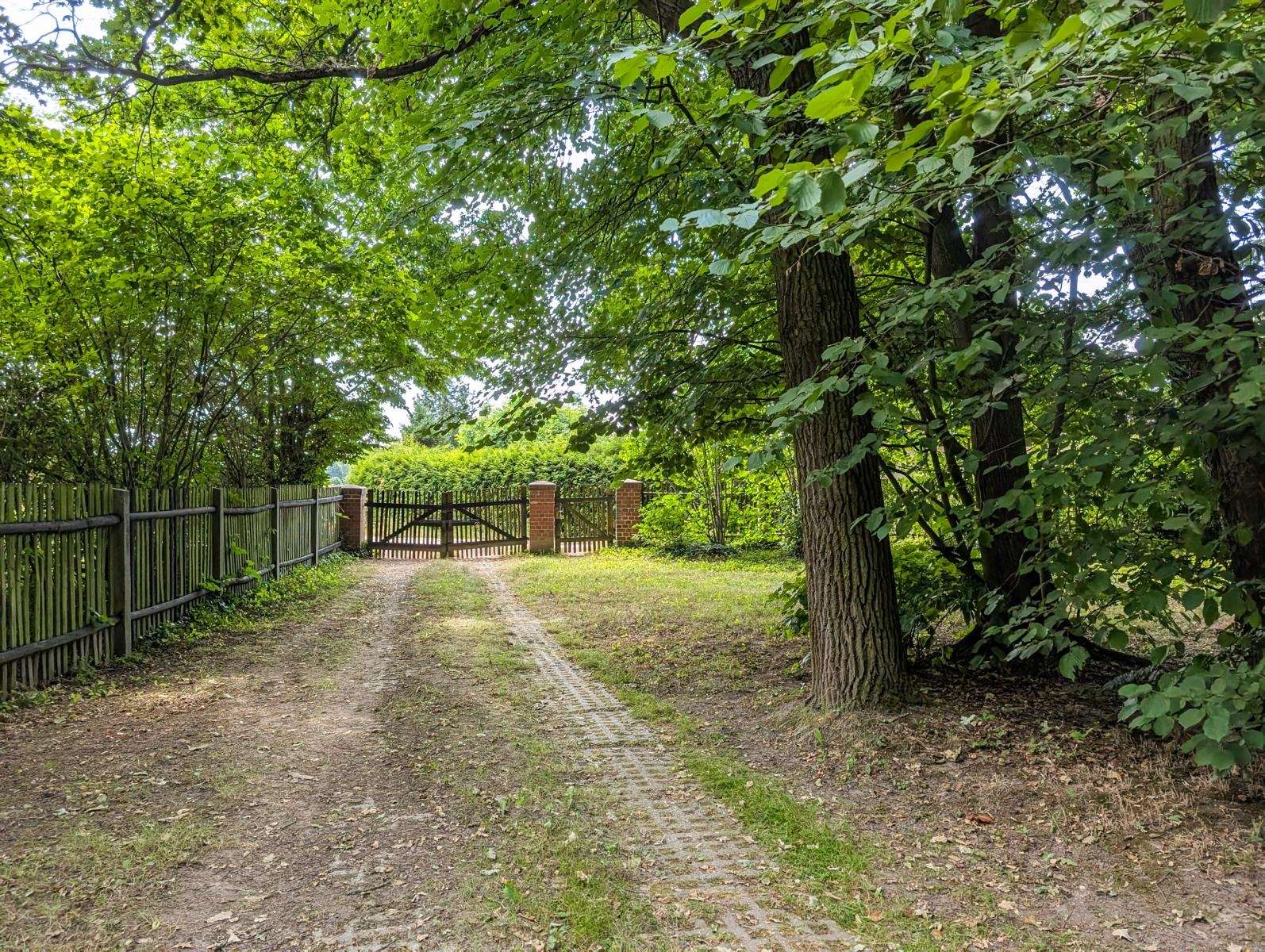 Dirt path with tire ruts leading to a wooden gate between brick posts, flanked by trees and a fence.