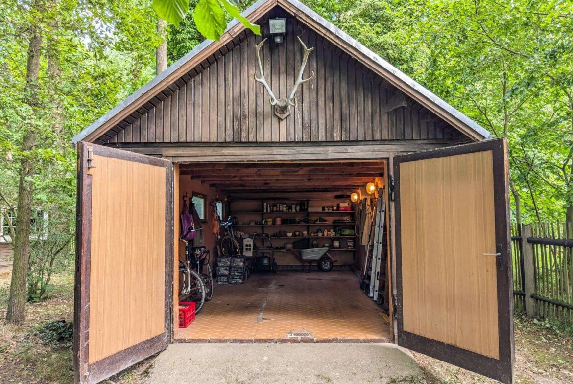 Wooden shed with antlers mounted on the peak, doors wide open to a cluttered workshop with shelves, tools, a bicycle, and a ladder inside.